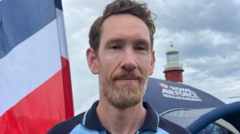A person wearing a light blue Royal Air Force Careers polo shirt stands in the foreground on Plymouth Hoe, with a large RAF-branded tent and a tall flag behind them. In the background, a red and white lighthouse and event displays are visible under a cloudy sky, with people moving around the Armed Forces Day site.
