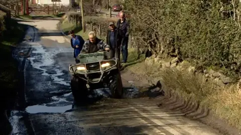 A man in a green four by four off-road vehicle navigating a pothole-ridden lane
