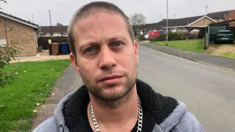 John Devine/BBC Lee Darling, 37, wears a gray woolen coat with black lining in the hood, a thick silver chain around his neck. He has a trimmed chin and short, dark hair, behind him is Papworth Road, where the attack took place, with bungalows on either side of the street.