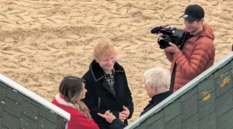 Amanda Jane Ed Sheeran - a man with ginger hair wearing a black jacket - standing on a beach next to a woman wearing a red jacket and a man with a camera and a cap. They are talking to an elderly man with grey hair.
