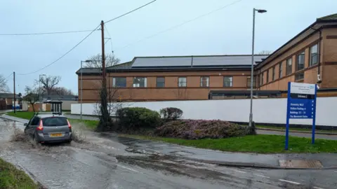 A grey car drives through floodwater on a road outside a hospital. There is a blue sign on the right hand side of the image, indicating it is an NHS site. There is a flower bed on a grass verge at the side of the road and brick buildings in the background.