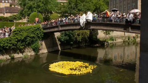 Bradford on Avon Town Council Large crowds of people on a stone bridge overlooking a river. They are emptying white plastic industrial bags, pouring thousands of yellow plastic ducks into the river below.