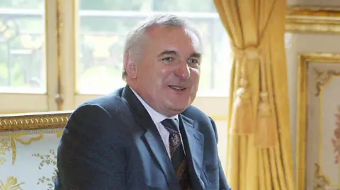 Getty Images An older men in a dark suit sits facing the camera in an ornate, gold-decorated room. He is seated on an elegant armchair smiling politely, with tall windows and gold curtains behind.