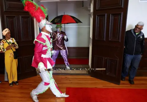Reuters A man wearing a colourful pink and white outfit wearing an elabourate hat posing and walking.