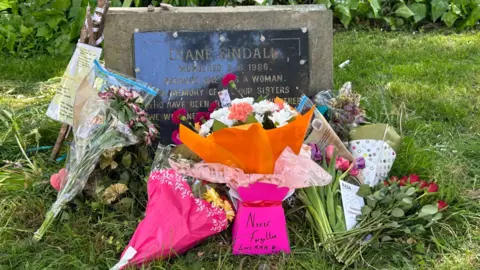 The grave stone of Diane Sindall, adorned with flowers. The inscription on it reads "murdered because she was a woman".