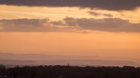 Getty Images The sun sets over the Somerset countryside with woodland and a tall structure pictured, as a silhouette, and rolling hills in the background.