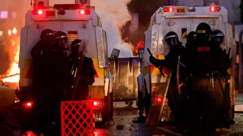 Riot police are gathered behind two large police Land Rovers - it is night time and in front of the two vehicles, behind which is a something on fire