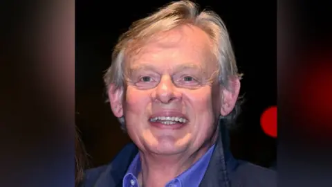 Martin Clunes seen in a picture, wearing a light blue shirt and a darker blue jacket 