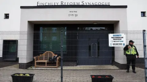 Getty Images/Toby Shepheard A photo of the front of Finchley Reform Synagogue, taken in mid-April after an arson attack. It shows a security fence and a security guard wearing a high vis yellow vest standing outside. The sign above the door is in Hebrew and then in English above it reads: Finchley Reform Synagogue. A sign on the security fence reads: SECURITY THESE PREMISES ARE PROTECTED BY SECURTEAM.