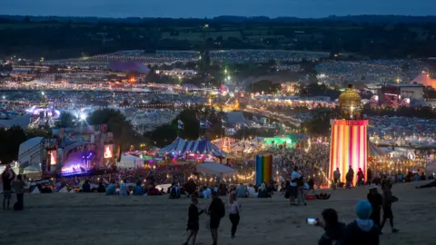 Getty Images A view of Glastonbury Festival from the signs, with the Park Stage and Ribbon Tower in shot. There are lights illuminating the dark evening sky, with festival goers filling the grassy spaces.