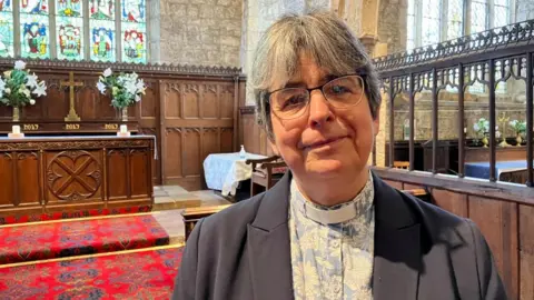 BBC/Seb Cheer A woman with short grey hair and wearing a light blue and white blouse with a flowery pattern stands in a church. She also wears a vicar's dog collar.