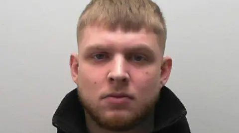 A pale faced man with short light coloured hair and a beard stares at the camera