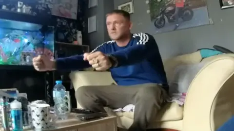 A man sat on a sofa with his arms in front of him. There is a photo behind him on the wall, a fish tank to his right and bottles and glasses on the table.