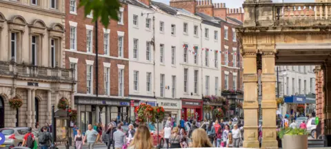 A busy town high street with people walking past shops and bunting hanging from the buildings 