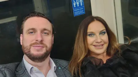 PA Media Photo of Pete and Emma Reid. Irish passengers who had planned to travel home via the storm-damaged Holyhead ferry port this Christmas. The couple are sat on the tube. Pete is wearing a checked suit jacket and stripey shirt, he has a beard and short brown hair. Emma is wearing a black chiffon blouse and has long brown hair. 