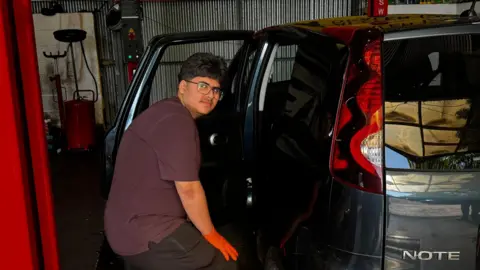 Kwick Fix Autocare Ltd Azhe is seen wearing orange gloves as works on a dark-coloured car inside a vehicle workshop. The rear door is open, with garage equipment and automotive service banners visible in the background.