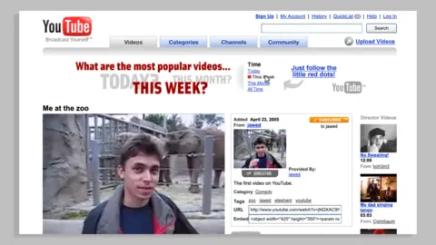 An early YouTube watchpage. The red and black YouTube logo is seen against a white web page background. The page features an image of Jawed Karim from his video 'Me at the Zoo'. Kawed is a young man with dark hair and is pictured standing in front of an elephant enclosure.