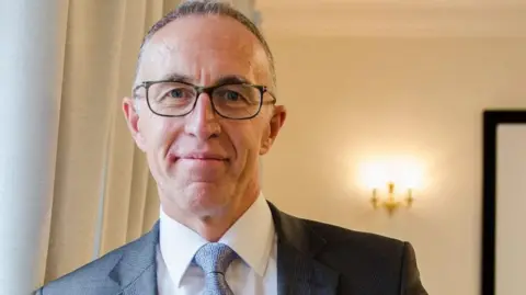 Lloyd Cooke wears glasses, a dark grey suit jacket, white shirt and pale blue paterned tie. He is smiling at the camera. 