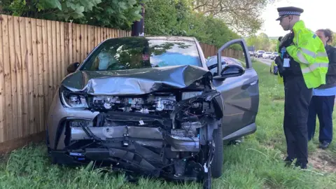A damaged car with a crushed front end has come to rest beside a wooden fence and grassy verge, while a police officer and a woman stand nearby assessing the scene along a roadside lined with trees.