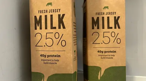BBC Two cartons of 2.5% fat reduced milk inside a fridge. The cartons are light brown and have writing on them including "MILK 2.5%" on them. There is also images of green cows on the cartons.