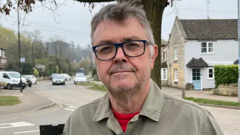 Martin Chillcott, 68, has brown hair with greying hair on each side. He is wearing a khaki shirt with an orange T-shirt under that. He has a large badge on the left of his shirt that says Protect Rural Peterborough. Roadways are visible behind him, with cars parked along near the kerbs. Some daffodils are in bloom on the left and a tree is behind him. There are some houses each side of the road.