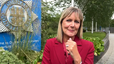 Joy has shoulder length blonde hair and a fringe. She is wearing a dark red trouser suit. She is pictured standing outside Durham police headquarters. 