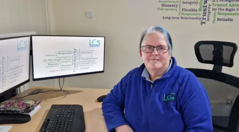 Emma Bygraves Emma Bygraves sat on a black desk chair behind a wooden desk with a computer, monitor, keyboard and notebooks on. The computer and monitor have a white screen with green and blue writing on. Emma has white and blue short hair and is wearing a bright blue fleece with a green logo on. She is sat in an office, the wall behind her is cream and has a patch of blue and green writing on.