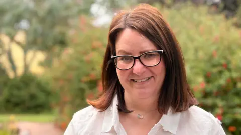 Anona Gauld is wearing a white shirt and black glasses. She has mid length brown hair and is smiling at the camera.