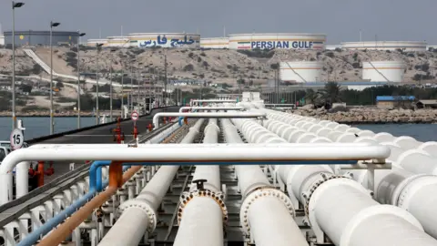 View of Kharg Island oil terminal, showing a row of pipes stretching into the distance towards a raised area of ground where several silos sit, one of them has the words "Persian Gulf" on its side.