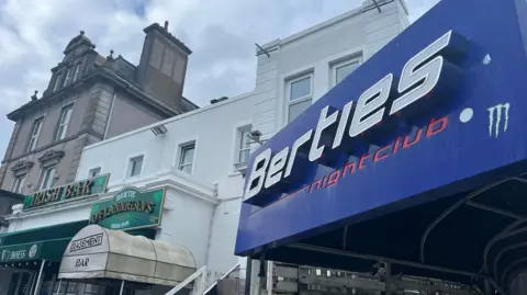 A large blue sign on the right reads Berties nightclub, while to the left is a pub with signage reading Bertie O'Flannigan's Irish Pub above a white building 