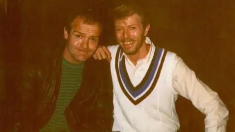 Supplied A photo of two men smiling at the camera. On the left is a man with short brown hair, a green top and leather jacket and on the right is singer David Bowie smiling with his hand resting on the man's shoulder. He is wearing a white V-neck jumper with blue, white and brown stripes.