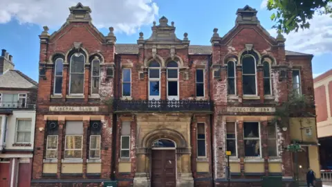 LDRS The Liberal Club a two storey red brick building with large windows and a balcony above the central doorway