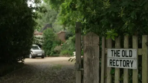 A sign on a wooden fence that is made out of white car number plates and reads "The Old Rectory". There is a driveway to the left of the fence and a car is parked at the bottom of the drive. 