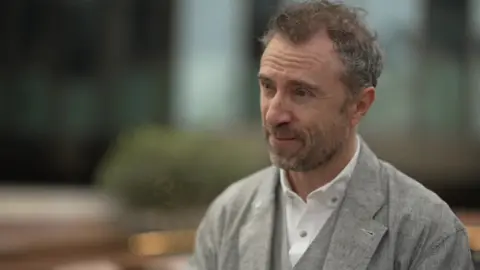 Close-up of a man in a grey jacket speaking during an interview. He stands against a blurred background.