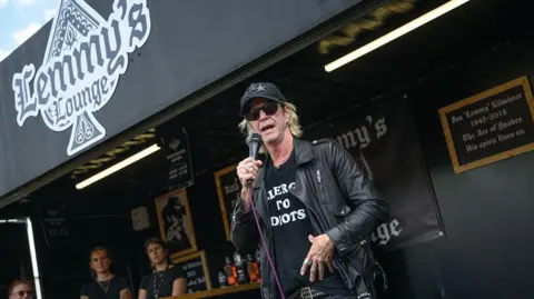 A man in a black T-shirt, a black leather jacket and a black cap with sunglasses speaks into a microphone. Behind him is a bar called "Lemmy's Lounge".
