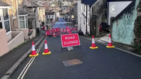 A red road closed sign has been put in the middle of a road in Bodmin with two traffic cones on either side of it. A set of red barriers and more traffic cones next to them have also been erected further down the road.