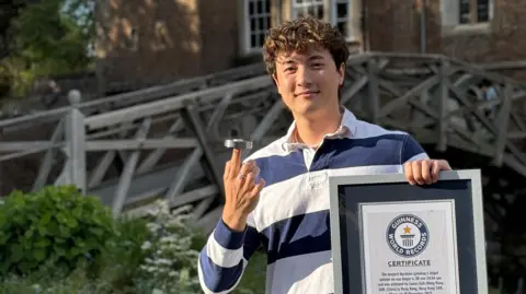 A young man is standing outdoors in front of a wooden, arched bridge that crosses a river, with an old college-style building visible behind it. He has short, curly brown hair and is wearing a blue-and-white striped rugby shirt. With his right hand, he is balancing a fidget spinner on one finger, demonstrating control and focus. In his left hand, he holds a Guinness World Record certificate. The setting has a calm, historic university atmosphere, likely on a campus.
