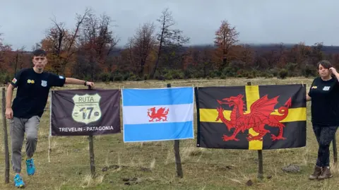 David Mardones David and Peace stand either side of three flags. One has the sign for a route 17, the other an Argentine flag with the Welsh dragon and the third is a Black flag with a yellow cross and a large red dragon.