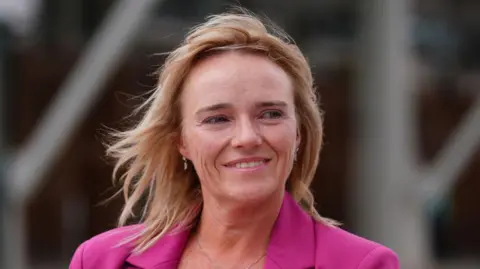 Nurse Sandie Peggie has short blonde hair and wears a pink blazer and smiles looking off to the side.