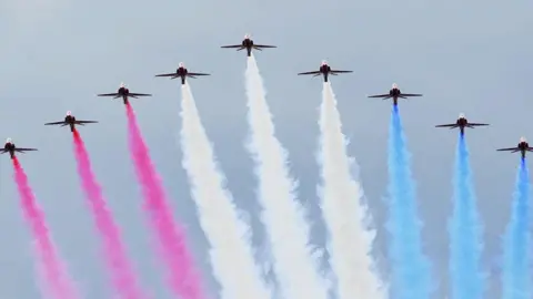 PA Media The Red Arrows during the flypast at RAF Fairford for the opening of the Royal International Air Tattoo. There are nine planes in the sky creating an arrow shape with red, white and blue smoke. 