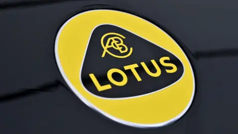 The Lotus logo. It has yellow writing on a black background surrounded by a triangular, curved shape. That is then surrounded by a yellow circle. The logo sits on a black, metallic surface.