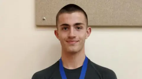 Jane Radford Will Radford is pictured smiling with a medal around his neck. He has short dark hair and wears a black t-shirt with a blue medal ribbon.