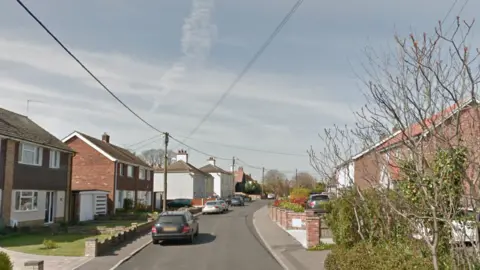 Google Maps A general view of Manor Road. Houses line the side of the road with vehicles parked up on the left hand side. Electricity cables travel over the road.