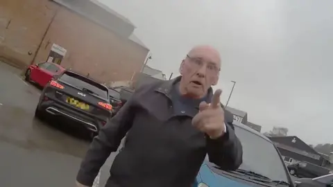 A man standing in a car park. He has his finger pointed at the person wearing the body-cam.