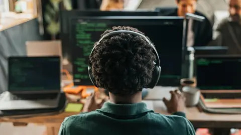 Getty Images A person, pictured from behind, sitting at a desk. There is a computer and two laptops in front of them. They have short brown hair and are wearing a dark green shirt and black headphones. 