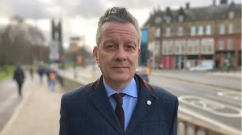 Nick Kemp, with short grey hair is standing on a high street. He is wearing a blue suit and is looking at the camera, expressionless.