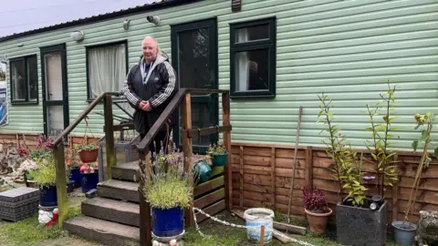 A woman with a black jacket standing outside a green static caravan