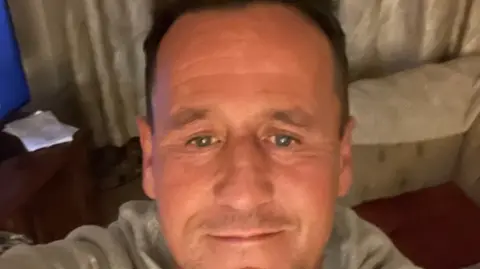 A selfie image showing a man in his 40s with receding brown hair. He is in a living room with a sofa and corner of a TV visible. He is wearing a light grey top. 