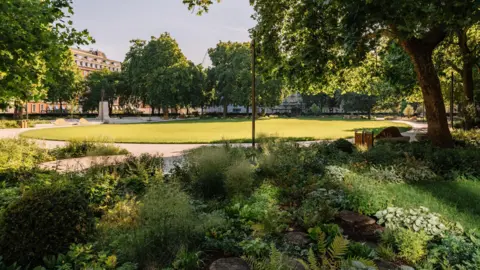Grosvenor An oval-shaped grass lawn bordered by a paved pathway. In the foreground, there is a dense planting of green ferns, shrubs, and low-lying vegetation. Mature trees cast shadows across the lawn, and a multi-storey brick building is visible in the background.