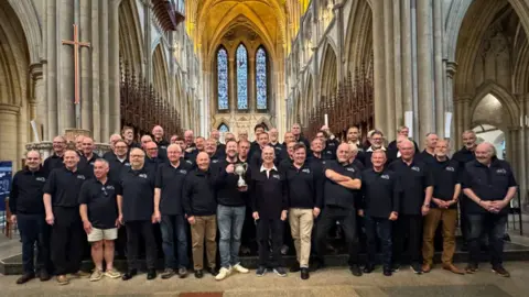 Bath Male Choir A group of men - mainly middle-aged and all dressed in blue polo shirts with the choir's logo - stand in the nave of a cathedral. They are all smiling to camera and one man in the middle is holding up a trophy. 